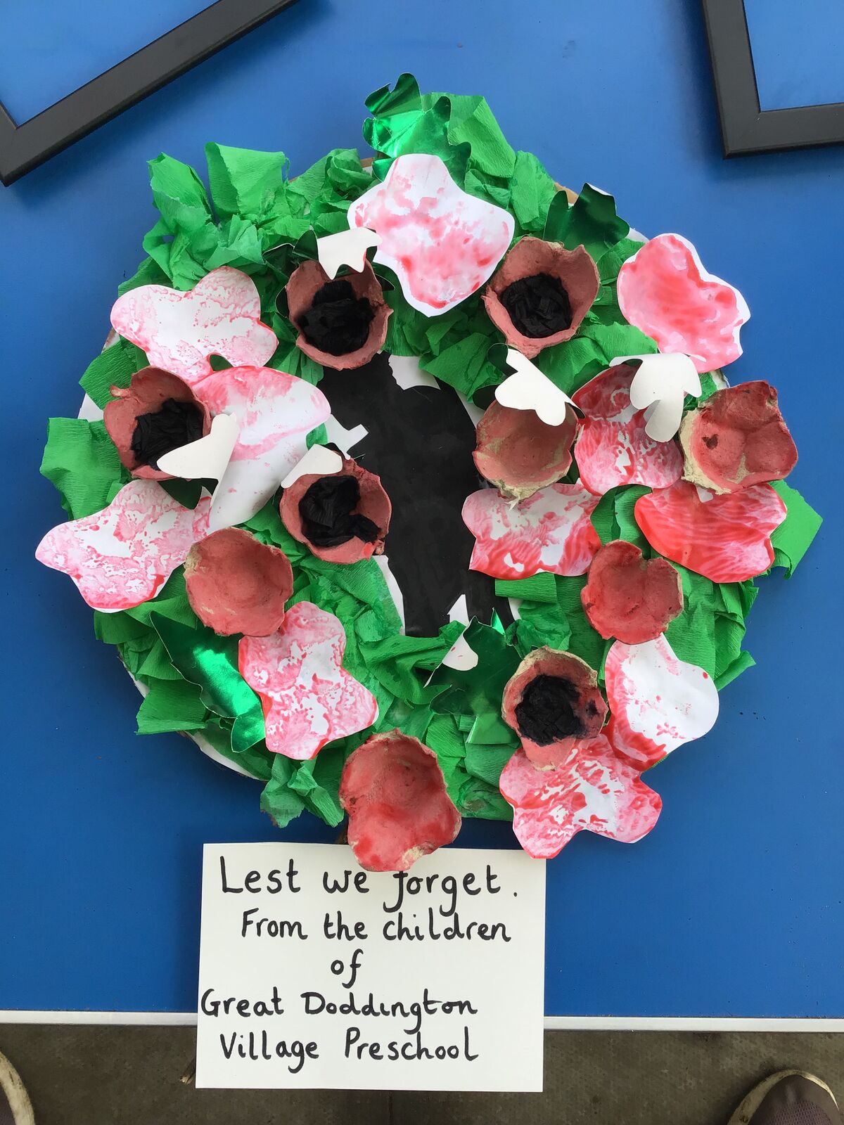 A handmade poppy wreath labelled 'Lest we forget, from the children of Great Doddington Village Preschool'