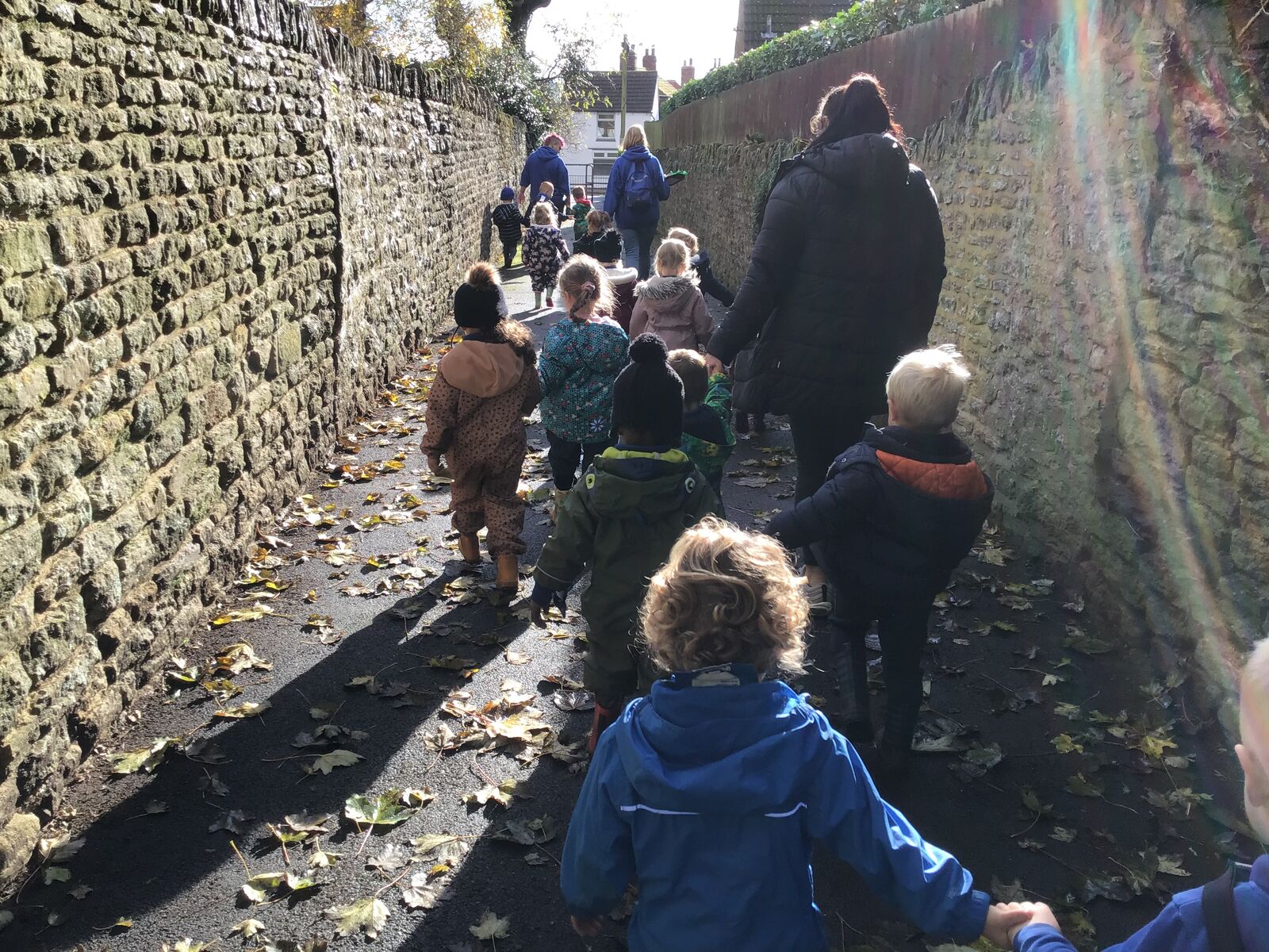 Children and staff walking together along a village lane beside a stone wall